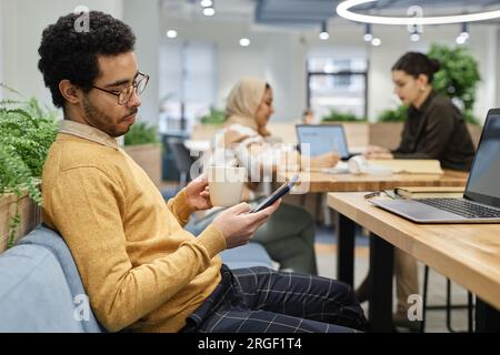 Side view portrait of young Middle Eastern man wearing glasses enjoying cup of coffee during break in office and checking messages, copy space Stock Photo