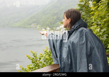 A young woman in a raincoat catches rainwater drops against the backdrop of a mountain lake and green hills Stock Photo