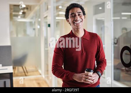 Waist up portrait of young Arabian man holding coffee cup while standing by glass wall in modern office and smiling at camera cheerfully, copy space Stock Photo