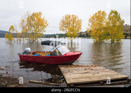 Red boat with outboard motor & canopy is tied to collapsed old dock on flooded, Lake Jindabyne in the Snowy Mountains of New South Wales in Australia. Stock Photo