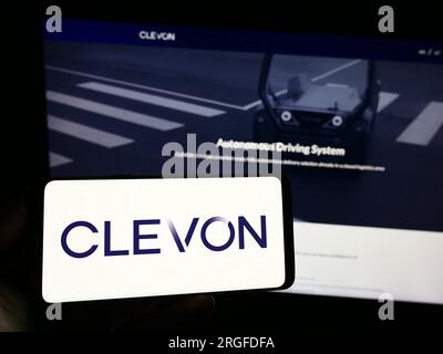 Person holding smartphone with logo of Estionian autonomous vehicle company Clevon AS on screen in front of website. Focus on phone display. Stock Photo