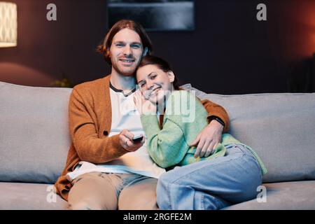 Front view portrait of smiling young couple watching TV at home and embracing, copy space Stock Photo
