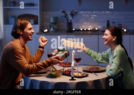 Side view portrait of happy young couple exchanging gifts while celebrating anniversary or Valentines day at home Stock Photo