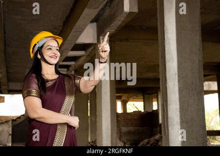 Portrait of confident young beautiful Indian female civil engineer or architect wearing saree and helmet holding pointing at something doing inspectio Stock Photo