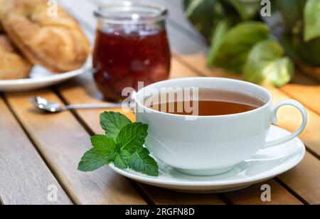 Cup of tea with fresh mint leaves and croissants and jar of jam in background on wooden patio table Stock Photo