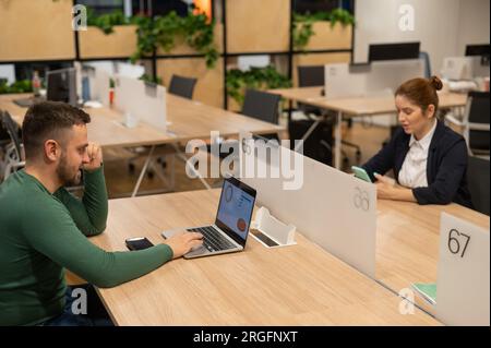 Red-haired caucasian woman uses a smartphone and a bearded man works behind a laptop in an open space office.  Stock Photo