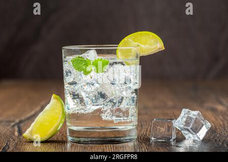 A glass of refreshing mineral water with ice cubes, mint leaves and lime slices on a wooden bar counter. Wooden background. Stock Photo