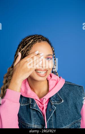 happy woman with dreadlocks covering eye and looking at camera on blue background, bold makeup Stock Photo