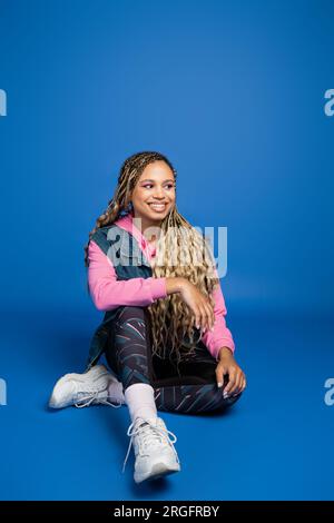 full length of young long haired african american woman sitting in sporty outfit on blue background Stock Photo