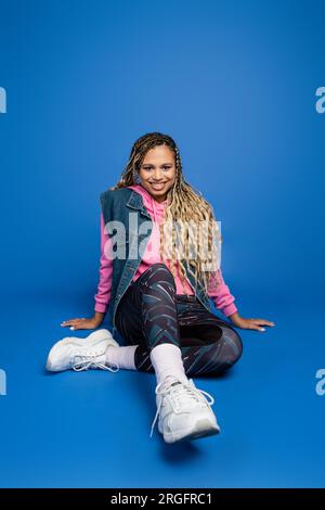 full length of joyful long haired african american woman sitting in sporty outfit on blue background Stock Photo