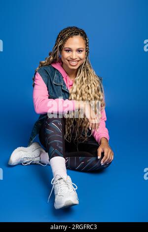 full length of happy long haired african american woman sitting in sporty outfit on blue background Stock Photo