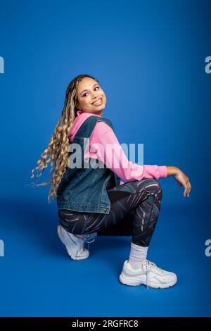 happy african american woman with dreadlocks sitting and posing on blue background, sporty outfit Stock Photo