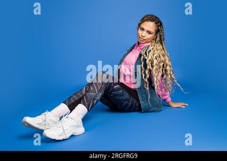 full length of long haired african american woman in sporty outfit sitting on blue background Stock Photo