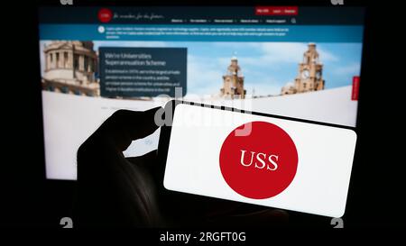 Person holding mobile phone with logo of Universities Superannuation Scheme (USS) on screen in front of company web page. Focus on phone display. Stock Photo