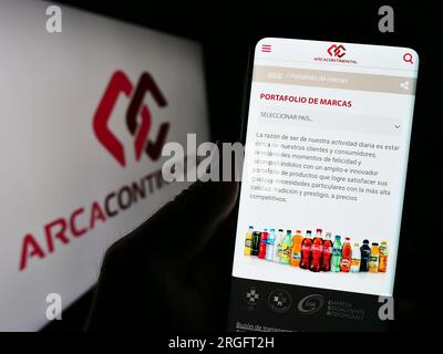 Person holding cellphone with webpage of company Arca Continental S.A.B. de C.V. on screen in front of logo. Focus on center of phone display. Stock Photo