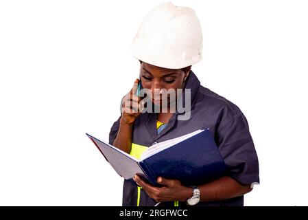 portrait of a beautiful female engineer wearing work uniform and white hard hat talking on phone and checking notes in notebook. Stock Photo