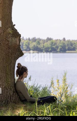 A captivating scene of enchantment, featuring a young brunette woman sitting against a majestic tree in the soft grass, her gaze fixed dreamily on the mesmerizing view of the forest lake. Lost in her thoughts, she finds solace in the tranquility of nature, allowing her dreams to take flight amidst the beauty and serenity of the natural world. Enchanted Dreams: Young Woman Gazing at the Forest Lake from Beneath a Tree. High quality photo Stock Photo