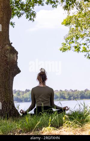 A serene and tranquil moment captured in nature, featuring a young brunette woman sitting gracefully in the lush green grass next to a majestic tree. She faces the peaceful forest lake, her eyes closed in deep meditation, as she embraces the soothing sights and sounds of the natural surroundings. This harmonious connection with nature creates a calming and rejuvenating experience for the soul. Serenity in Nature: Young Woman Meditating by the Forest Lake. High quality photo Stock Photo