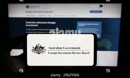 Person holding cellphone with logo of Australian Foreign Investment Review Board (FIRB) on screen in front of webpage. Focus on phone display. Stock Photo