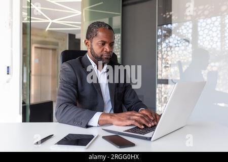 Serious mature businessman working on laptop inside office at workplace, african american boss thoughtful in business suit, successful experienced investor banker. Stock Photo