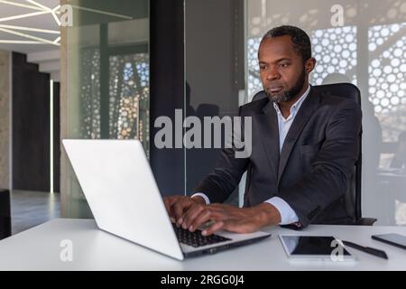 Serious mature businessman working on laptop inside office at workplace, african american boss thoughtful in business suit, successful experienced investor banker. Stock Photo
