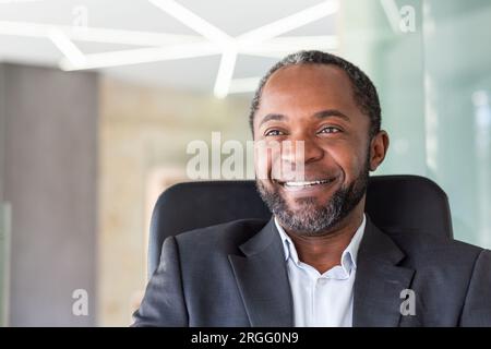 Close up of successful dreamy businessman smiling and looking at camera, african american boss in business suit smiling and looking at window inside office. Stock Photo