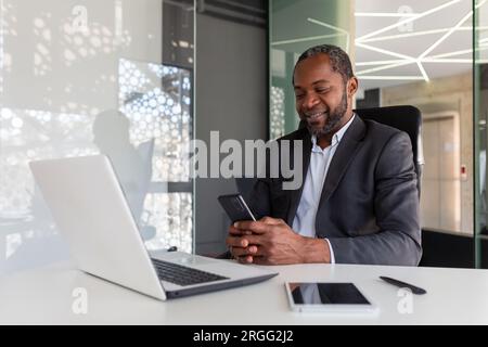 Mature experienced and successful african american boss at workplace inside office, senior satisfied with results businessman using phone, reading news from online app on smartphone. Stock Photo