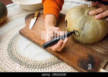 top view of female hands of unrecognizable person, drawing with black marking eye on pumpkin on wooden board, which she is decorating for halloween, c Stock Photo