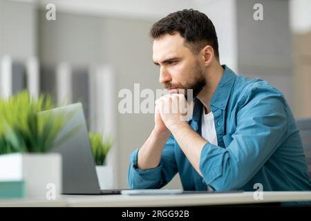 Pensive Young Businessman Sitting At Workplace And Looking At Laptop Screen Stock Photo
