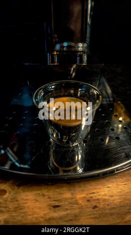 Modern design glass cup of glass with freshly brewed black coffee with golden foam with small bubbles on top of an automatic barista espresso machine Stock Photo