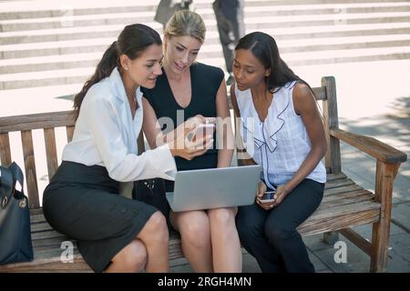 Colleagues using smart phone and laptop on bench Stock Photo