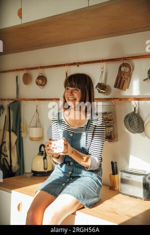 Woman sitting on kitchen bench holding coffee cup Stock Photo