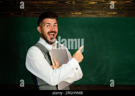 Back to school. Happy teacher near chalkboard pointing at copy space. Smiling student with laptop in classroom. Education, e-learning, teaching. Male Stock Photo