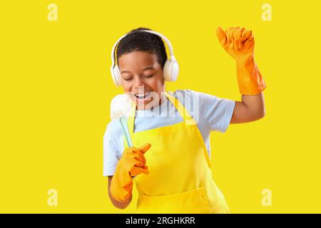 Cheerful African-American boy in headphones singing and using brush as microphone on yellow background Stock Photo