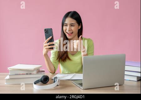 A positive and pretty Asian girl shows her thumb up and enjoys talking on video call with her friend while sitting at her study table against an isola Stock Photo