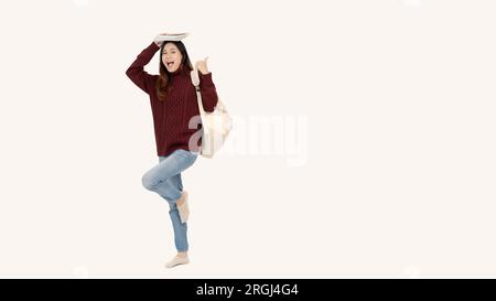 A joyful and pretty young Asian female college student in a red sweater holds books over her head and poses in a cute full-body pose against an isolat Stock Photo