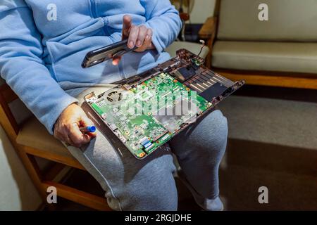 computer repairs, old woman in her seventies repairing her laptop, holding the phone light to see better Stock Photo