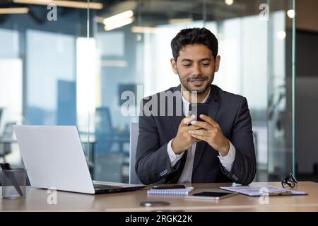 Successful experienced businessman using phone while sitting at workplace, hispanic smiling happy with achievement results holding phone, reading online using app on smartphone. Stock Photo