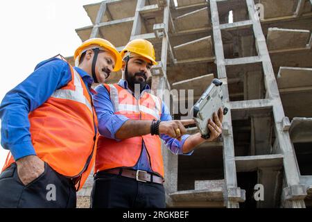 Two Indian male civil engineers or architect wearing helmet and vest holding digital tablet blueprint at construction site discussing real estate proj Stock Photo