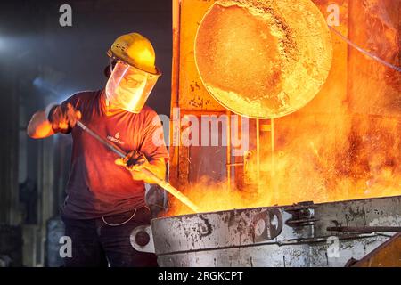 factory worker with helmet and safety gear steering a big pot of melted metals. machine pouring liquid metals into a mold Stock Photo