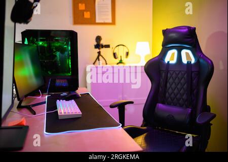 Modern gadgets computer monitor connected keyboard neon lights gaming PC video camera headphones and cozy headrest armchair placed in illuminated room Stock Photo