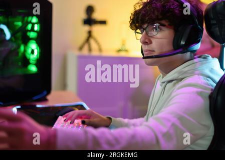 Side view of concentrated young male gamer in eyeglasses headphones looking at screen of monitor while sitting at table and playing on keyboard in roo Stock Photo