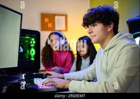 Side view of focused young male gamer looking at screen of monitor while sitting at table and playing with keyboard of computer with diverse girlfrien Stock Photo