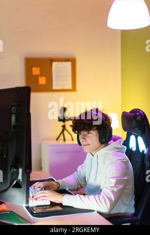 Side view of concentrated young male gamer headphones looking at screen of monitor while sitting at table and playing on keyboard in room Stock Photo
