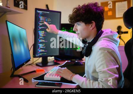Side view of focused young male looking at screen of monitor with friends in conference while sitting at desk and pointing finger at program on screen Stock Photo