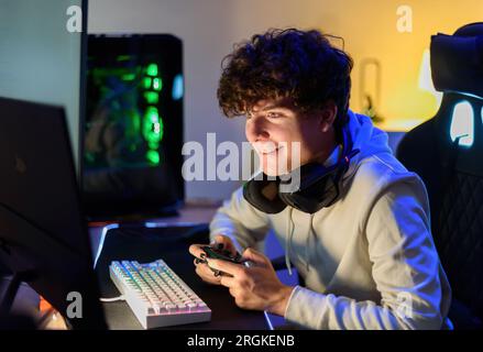 Focused smiling young male looking at screen of computer while sitting at table with gaming PC and playing video games alone with joystick in room Stock Photo