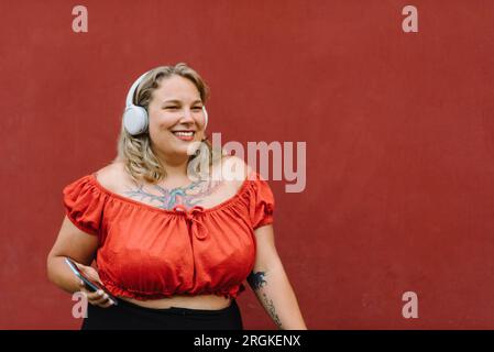 Positive body young female in trendy outfit and headphones smiling and looking away while standing on red background and enjoying music Stock Photo