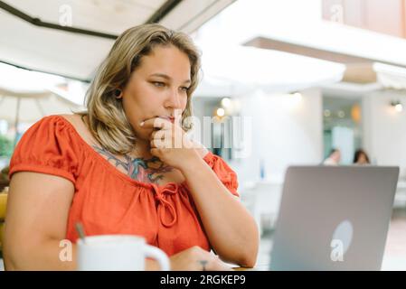 Portrait of pensive chubby young female freelancer in casual clothes sitting at table with laptop and cup of hot drink in cafe while working outdoors Stock Photo
