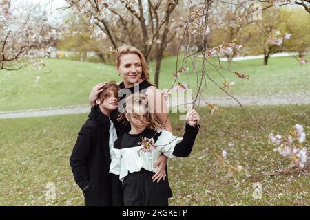 Happy woman and children in black outfits standing under blooming tree in park and embracing Stock Photo