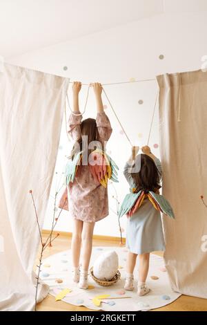 Full body back view of anonymous little girls in casual clothes and mini colorful wings standing with raised hands and holding rope of curtain while p Stock Photo